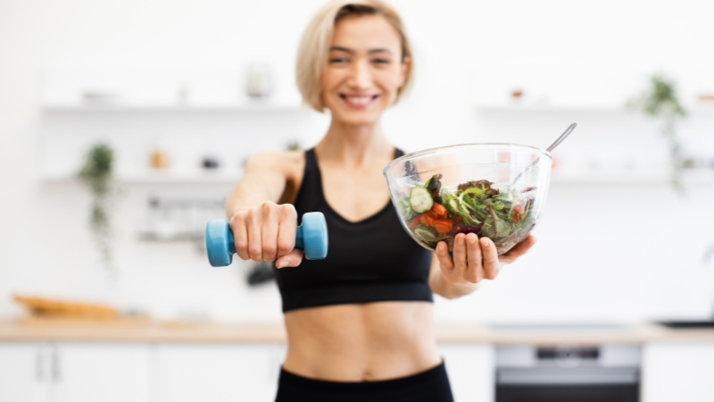 Nasmejana žena u sportskoj opremi drži teg u jednoj ruci i činiju salate u drugoj